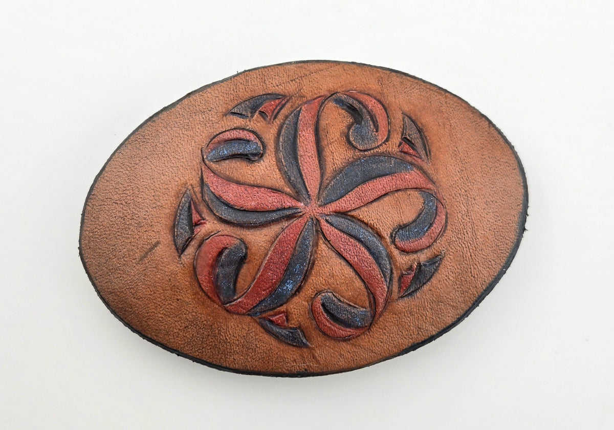 Front of the Hand Tooled Leather Vintage Belt Buckle — Oval Red & Navy Pinwheel, showing an oval face with a red and navy pinwheel floral scroll on tan leather.