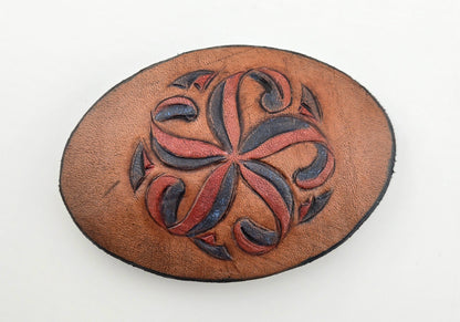 Front of the Hand Tooled Leather Vintage Belt Buckle — Oval Red & Navy Pinwheel, showing an oval face with a red and navy pinwheel floral scroll on tan leather.
