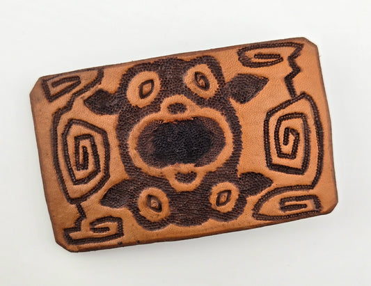 Front of the Hand Tooled Leather Vintage Belt Buckle — Geometric Tribal Spirals, showing a bold geometric tribal design of dark carved spirals and shapes on a light tan leather face.
