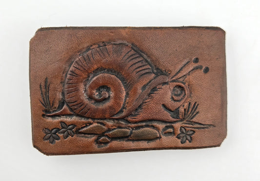 Front of the Hand Tooled Leather Vintage Belt Buckle — Snail with Spiral Shell, showing a large snail with a deeply carved spiral shell, flanked by small flowers and grass blades on tan-brown leather.